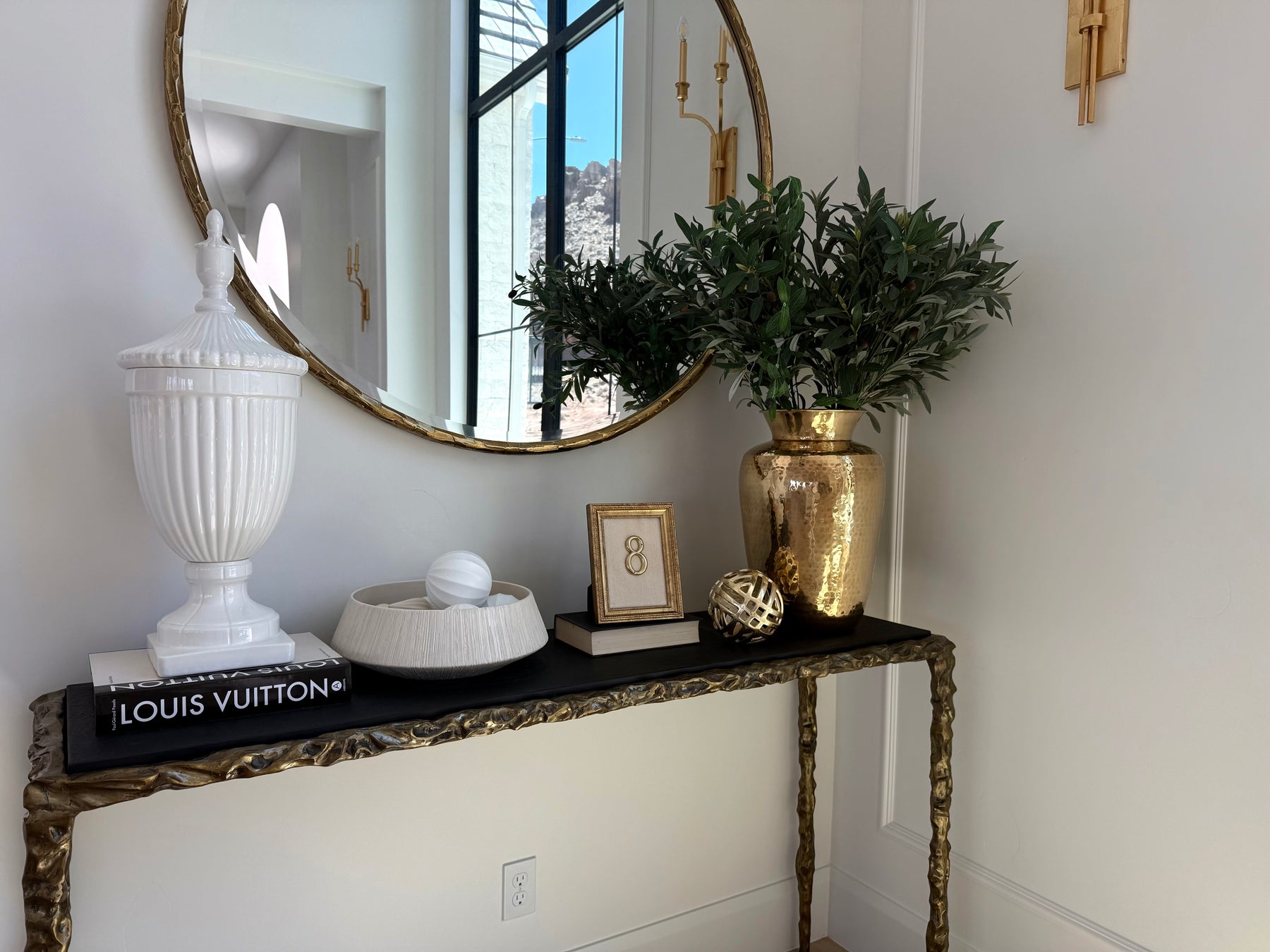 A styled interior corner featuring a gold-framed oval mirror, a black console table with gold legs, a white ribbed urn decor piece, a white bowl, a gold vase with green foliage, and a Louis Vuitton book on a white wall.