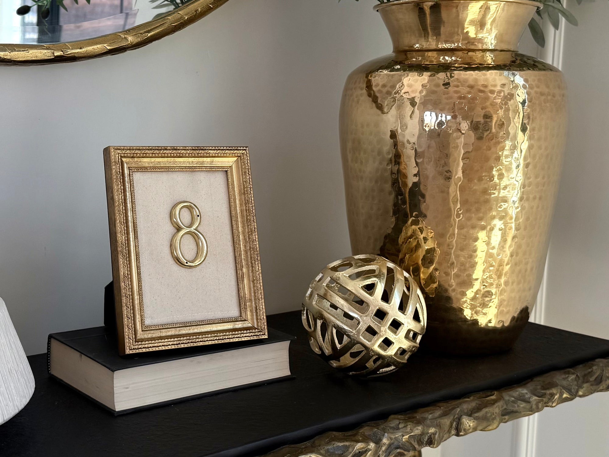 A styled console table featuring a large hammered gold vase filled with green foliage, a black tabletop, a gold-framed picture of the number 8, a white textured bowl, and decorative gold and white spheres.
